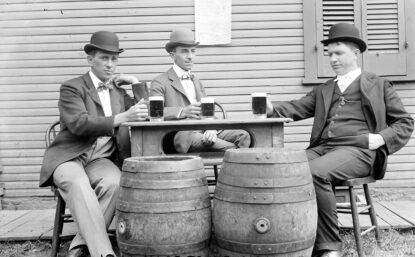 Black and white photograph of men in suits sitting around table holding beers with two barrels in front. Writing on photo says "Nice to have been waited on."