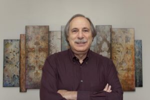 Bernard Lightman, dark red collared shirt, arms folded, in front of an artwork on a wall