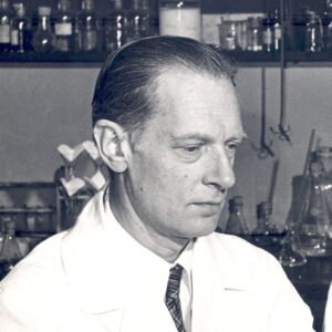 Carl and Gerty Cori in their lab, wearing white lab coats. Carl points to a document with a pencil. There are beakers and bottles in the background.