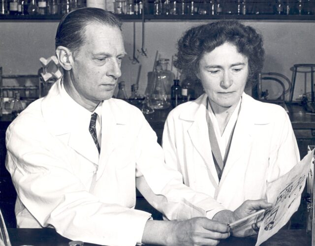 Carl and Gerty Cori in their lab, wearing white lab coats. Carl points to a document with a pencil. There are beakers and bottles in the background.