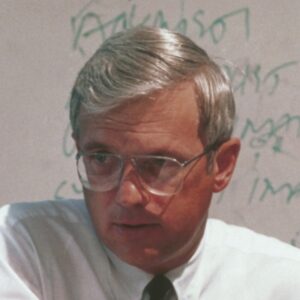 Fuller in a white button up and tie, wearing glasses and sitting in front of a dry erase board