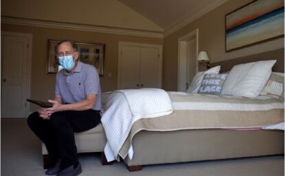 Photograph of Robert Langer sitting at the foot of a bed, wearing a surgical mask.