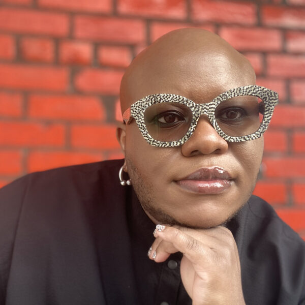 Cecilio in a black dress-shirt, leopard-print glasses, resting head on hand, in front of a bright red brick wall