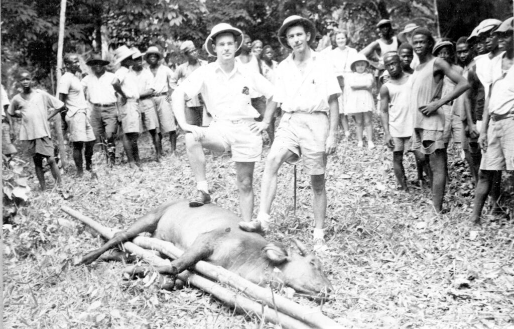 Two men in pith helmets standing with their feet on a dead animal while a large group looks on