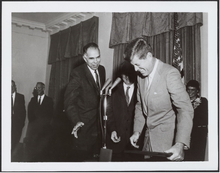 Glenn Seaborg and President John F. Kennedy present the AEC’s Enrico Fermi award to Dr. Hans Bethe, December 1, 1961. Seaborg in a suit extends his right hand while Kennedy, in a suit, laughs and stands at a podium with 2 microphones.