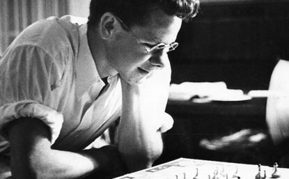 Black and white photograph of a young Gordon Gould looking down at a machine. He is wearing glasses and a white button down shirt.