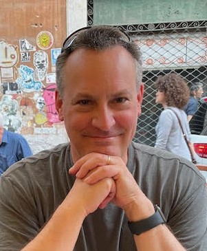 John Powers outdoors in an urban area, wearing t-shirt, resting chin on hands