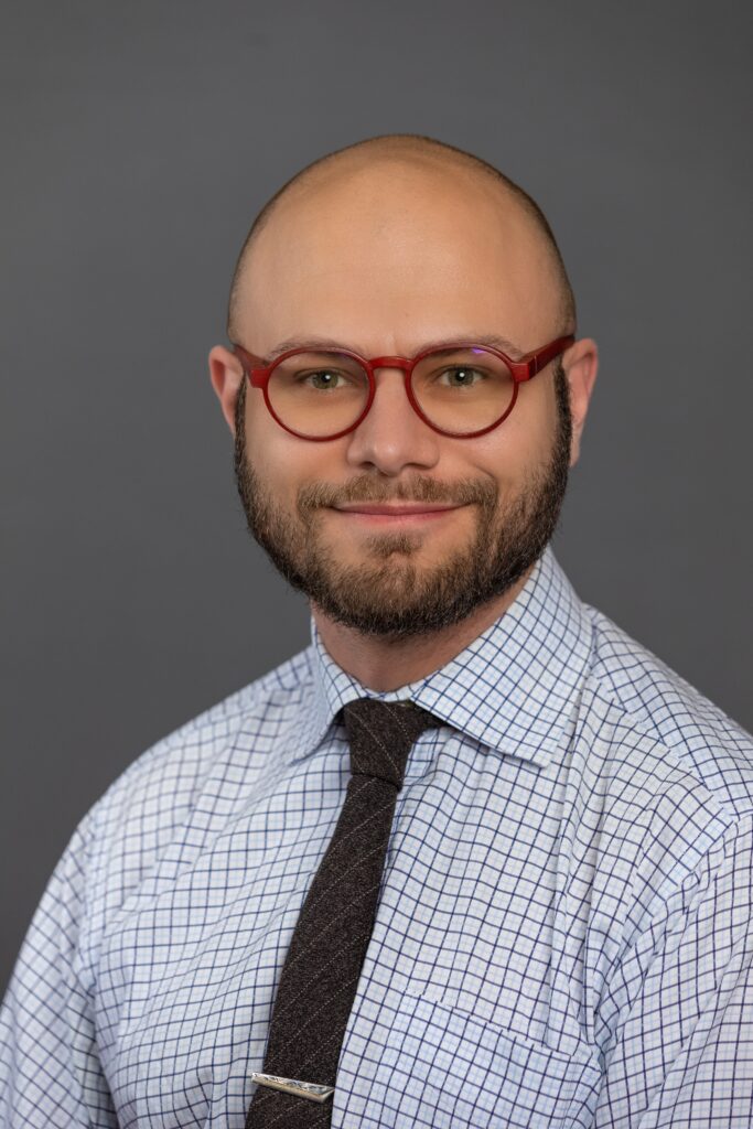 Julian Silverman with gray studio backdrop, red frame glasses, beard, smiling, checkered shirt, tie with clip