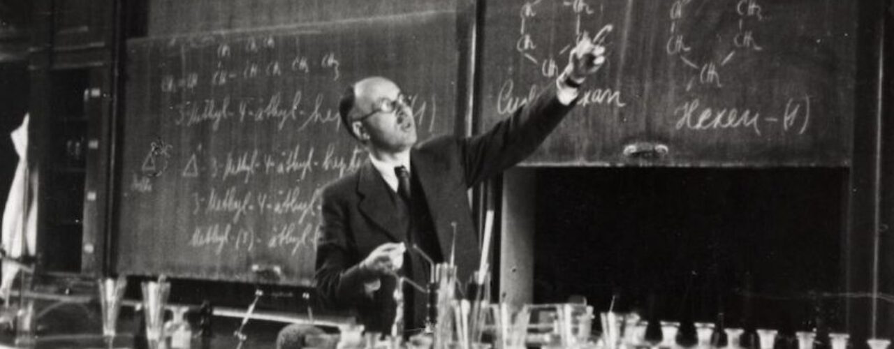 A black and white photo of a man wearing glasses in a suite and tie pointing at a chalk board. There is a counter filled with glass vessels in the foreground.