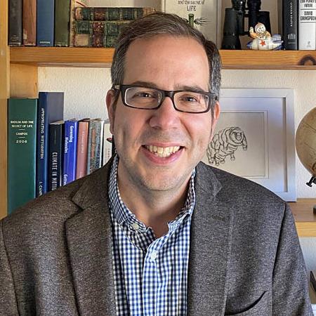 Luis Campos smiling, wearing glasses, in front of bookshelf