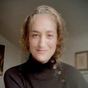 Mariel wears a black turtle neck with buttons on the side. Her curly hair is pulled back.