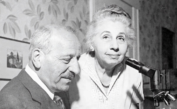 Black and white photo of a seated older man and standing older woman in front of a microscope