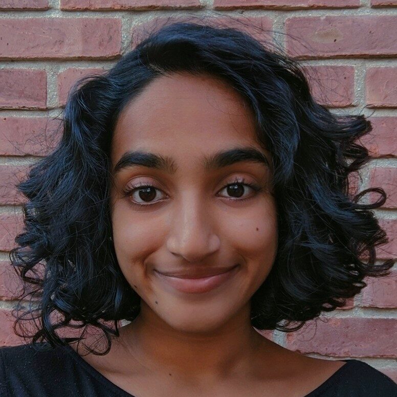 Nandini smiling wearing their short curly hair down. There is a brick wall behind them.