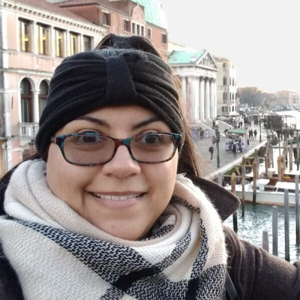 Nyomi smiles and wears a thick black headband and plaid scarf. There is a canal in the background.