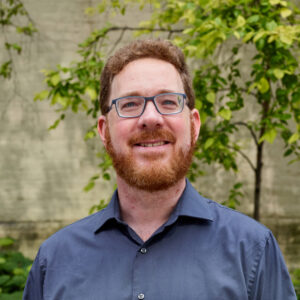 Roger outdoors, looking up and smiling, wearing glasses and blue button-down shirt