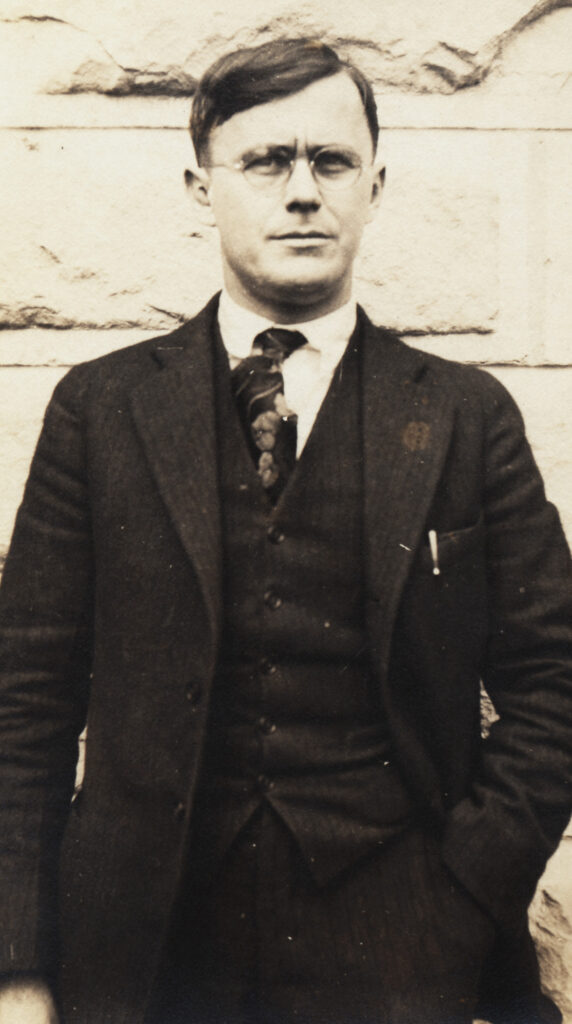 Black and white photo of a standing man in glasses and a suit