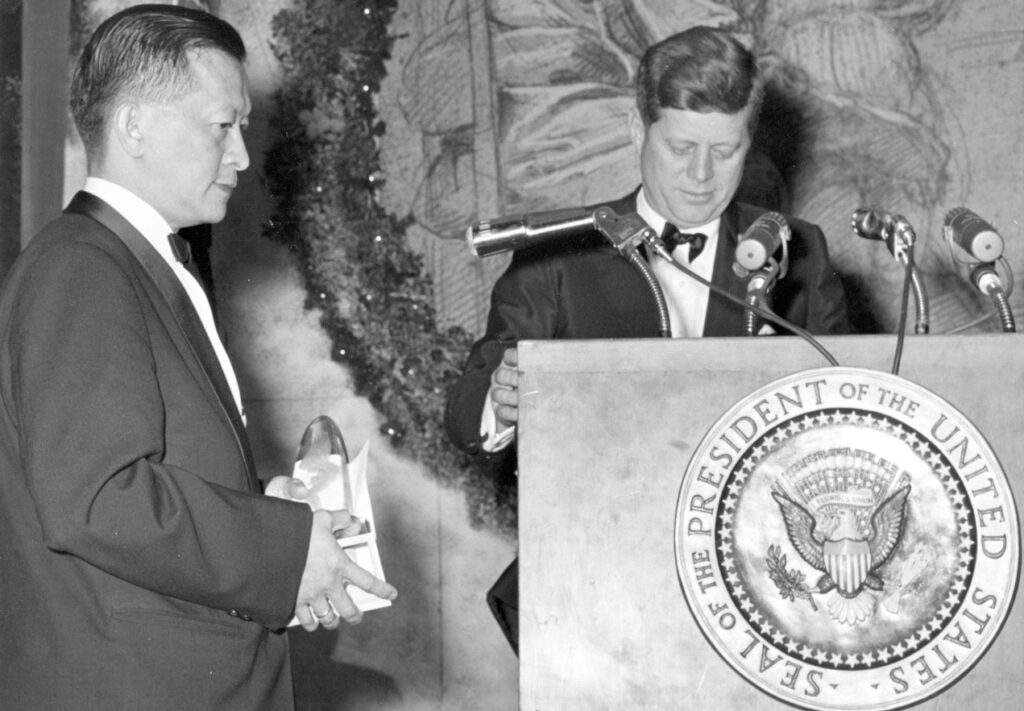 Black and white photo of two men in tuxedos, one standing behind a lectern with a seal and microphones, the other holding an award