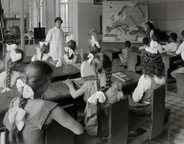 1920s black and white photograph of children learning geography in a classroom. Girls and boys are seated on different sides of the room.