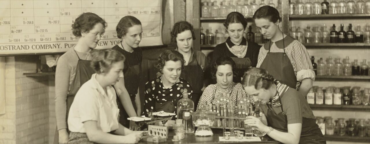 group of women standing around a lab table with periodic table in background