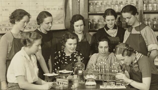 group of women standing around a lab table with periodic table in background